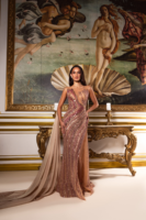 Model posing in an elegant floor-length evening gown with sheer illusion neckline, intricate sequin embellishments in gold and pink tones, high side slit, and flowing champagne chiffon train, set against a backdrop featuring Botticelli's *The Birth of Venus*.
