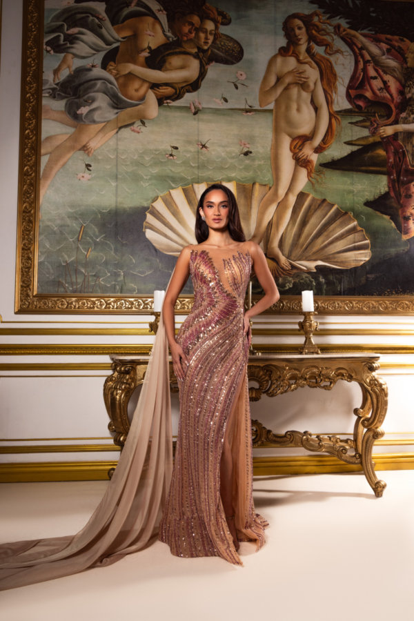 Model posing in an elegant floor-length evening gown with sheer illusion neckline, intricate sequin embellishments in gold and pink tones, high side slit, and flowing champagne chiffon train, set against a backdrop featuring Botticelli's *The Birth of Venus*.