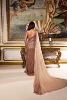 Model wearing a floor-length, one-shoulder sequin gown in rose gold with vertical stripes, featuring a flowing nude chiffon cape train, shown from the back in an ornate setting with a classical painting.
