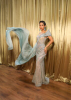 Elegant floor-length off-shoulder evening gown in shimmering light blue-silver sequins with intricate embellishments, sheer panels, and a flowing chiffon train dramatically swept to the side. Model poses confidently against golden curtains.