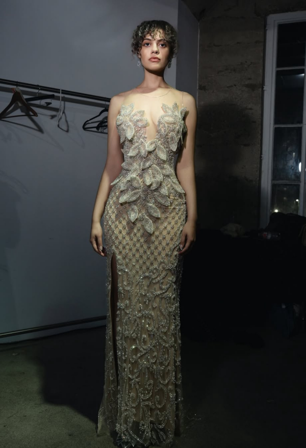 Elegant floor-length evening gown in shimmering nude with intricate silver beadwork, featuring sheer illusion neckline, large floral appliqués on the bodice, fitted silhouette, and high side slit.