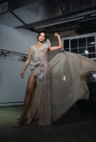 Elegant sheer champagne evening gown with intricate sequin embellishments, high side slit, dramatic ruffled shoulder detail, and flowing train, modeled in a dynamic pose.