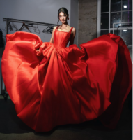 Model wearing a vibrant red satin ball gown with a structured corset bodice and voluminous full skirt, twirling to show the dramatic flow of the fabric.