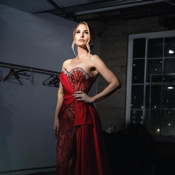 Elegant strapless red evening gown with intricate sequin and bead embellishments on the bodice, asymmetrical draped satin overlay, and high side slit, modeled in a dimly lit studio.