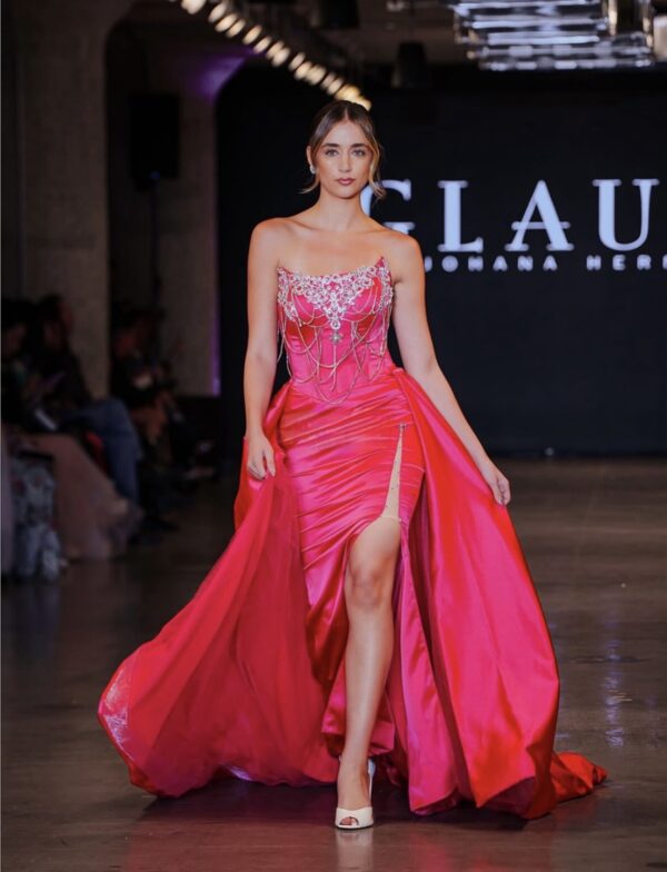 Model walking down the runway in a vibrant pink strapless evening gown with intricate silver embroidery, beaded chains, high side slit, and flowing train.