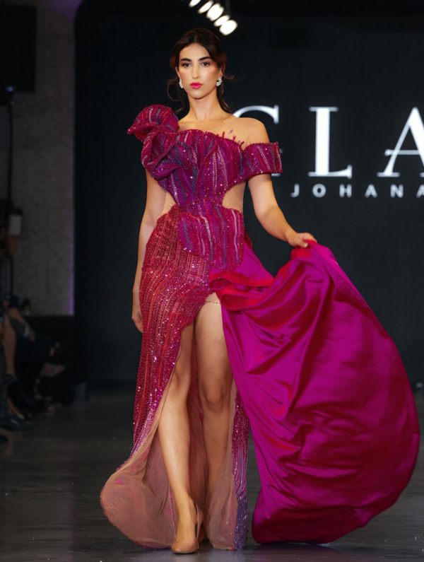 Model wearing an elegant off-shoulder magenta sequined evening gown with dramatic ruffled shoulder detail, side cutout, high slit, and flowing train on the runway.
