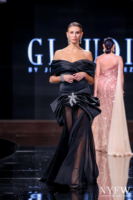 Model on the runway wearing an elegant off-the-shoulder black evening gown with dramatic draped bodice, ruched peplum, sparkling crystal bow embellishment, and sheer flowing skirt overlay.